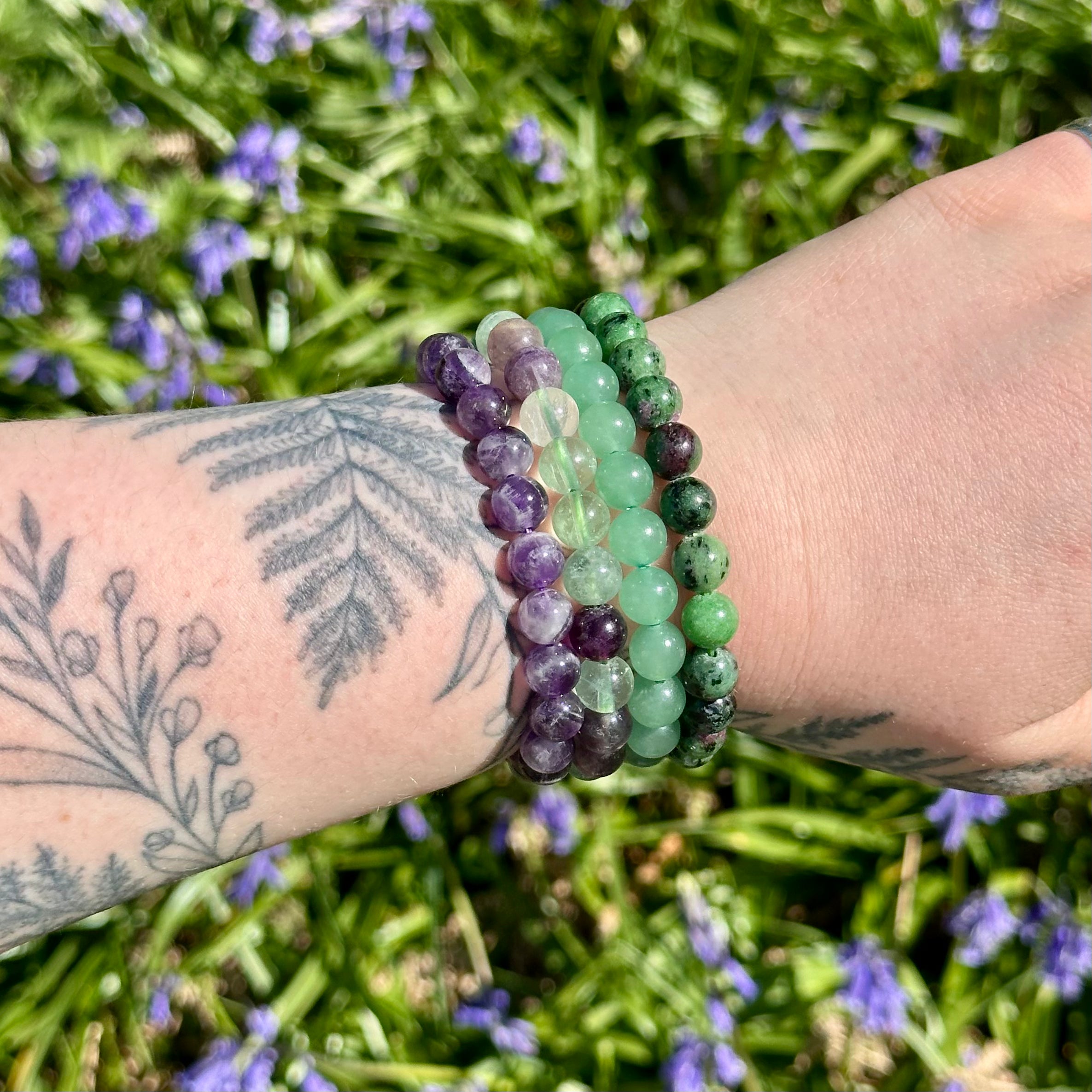Stack of beaded bracelets on a wrist with tattoos, against a natural background. Amethyst, Fluorite, Green Aventurine and Zuby Zoisite 