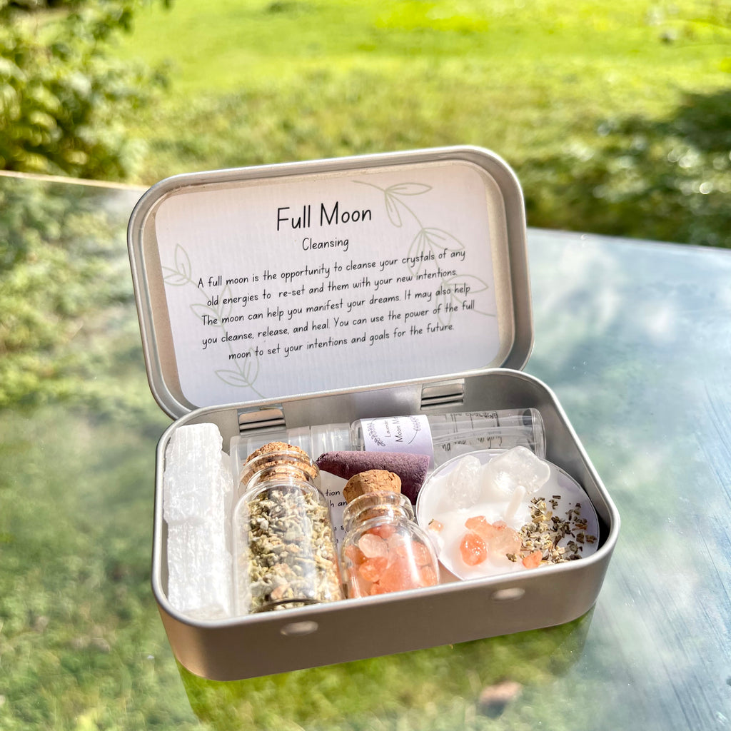 A travel altar tin opened to display its contents including jars of sage and salt, a selenite stick, a clear quartz infused lavender moon water, and a lavender scented incense cone, all arranged neatly inside.