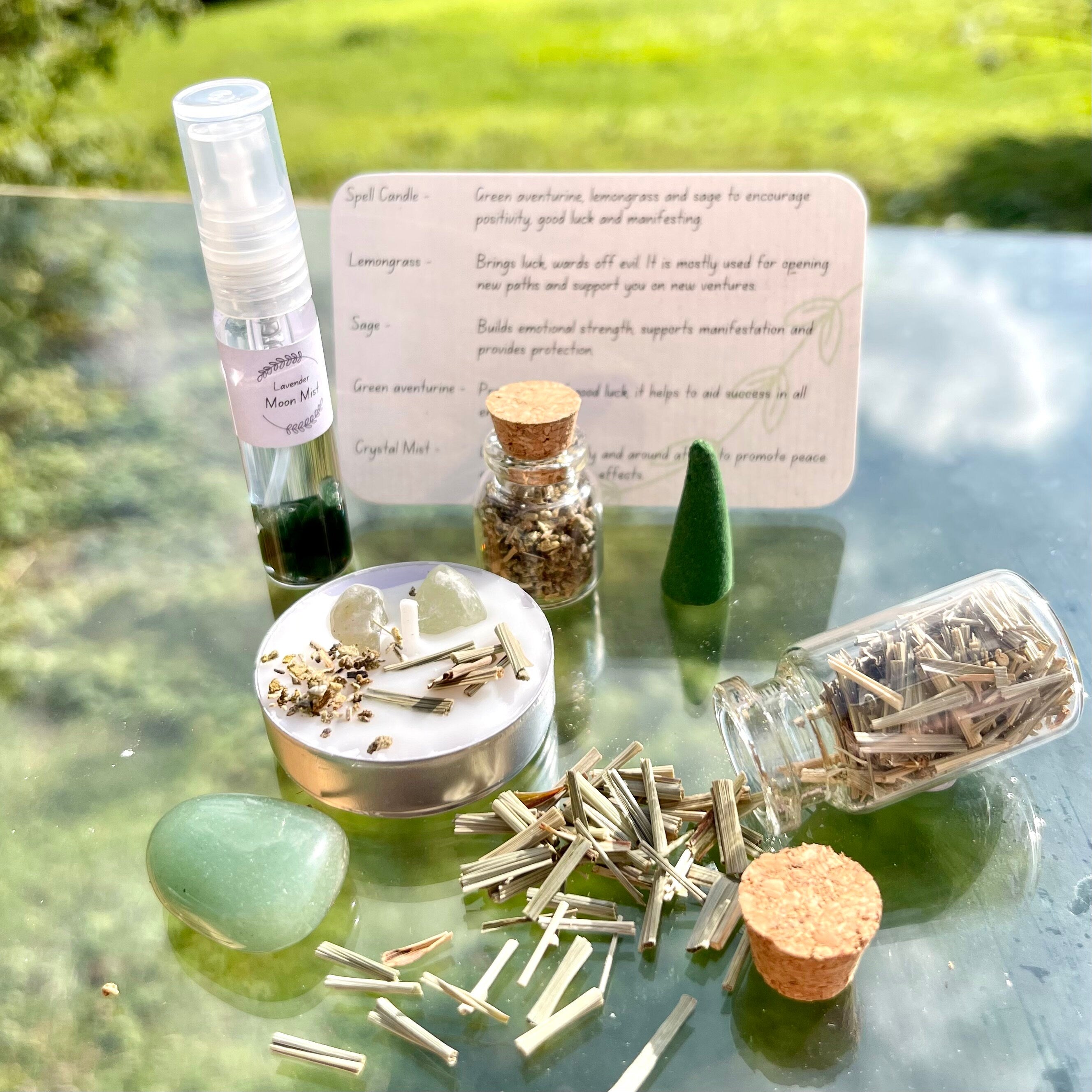 The contents of the good luck travel altar laid out. A moon mist with green aventurine crystal chips, a jar of sage, jar of lemongrass, spell candle, incense cone and green aventurine tumblestone.