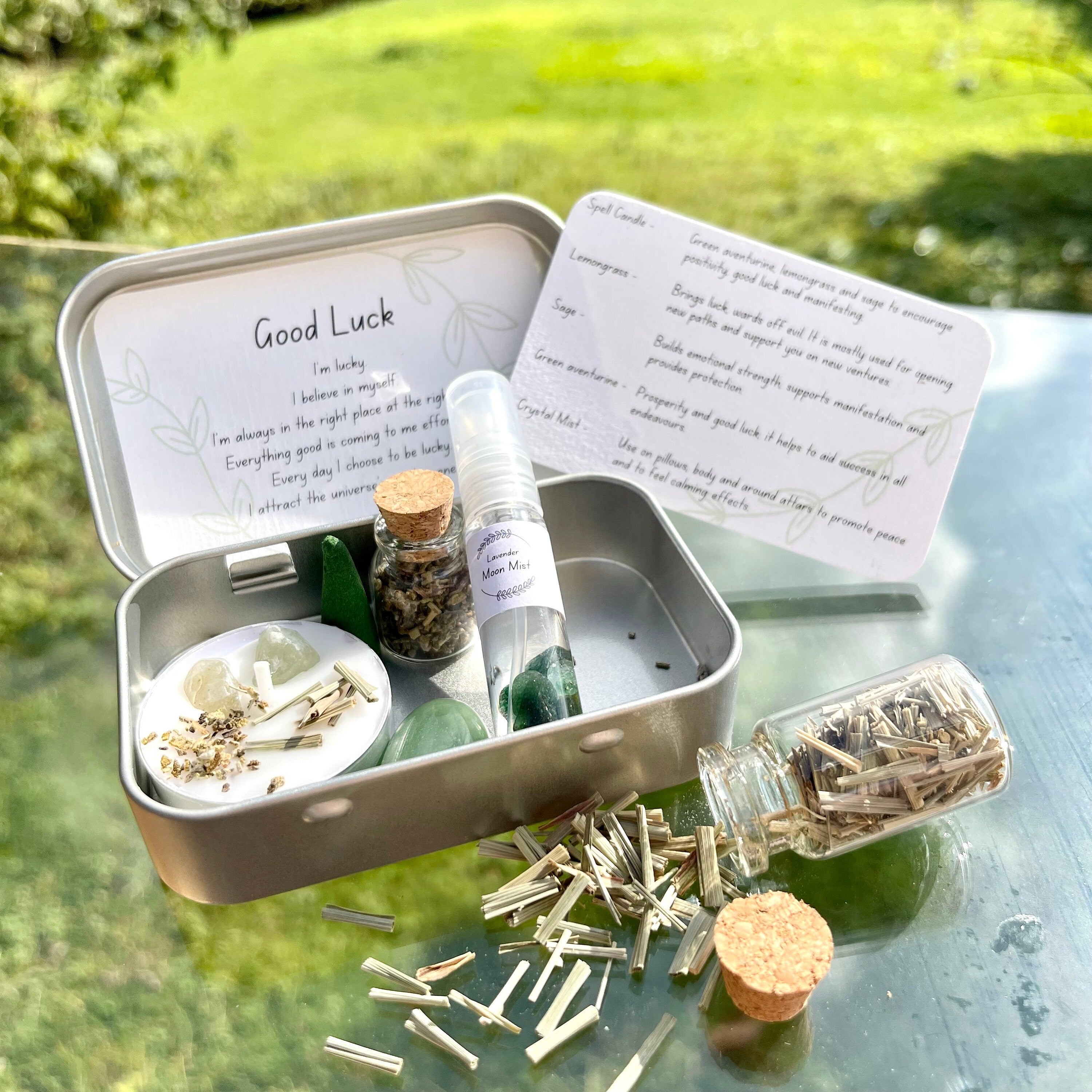 A miniature Good Luck travel altar tin opened to display its contents including a white spell candle with herbs, jars of dried herbs, a green stone, a moon mist, and incense cone, green aventurine tumblestone, all set on a glass surface with a backdrop of grass.