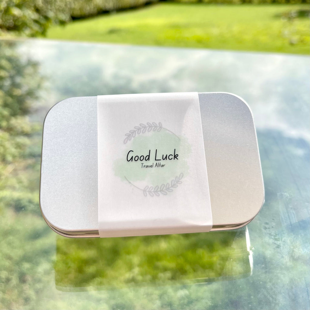 A metal tin with paper wrap with a green good luck travel altar logo on it