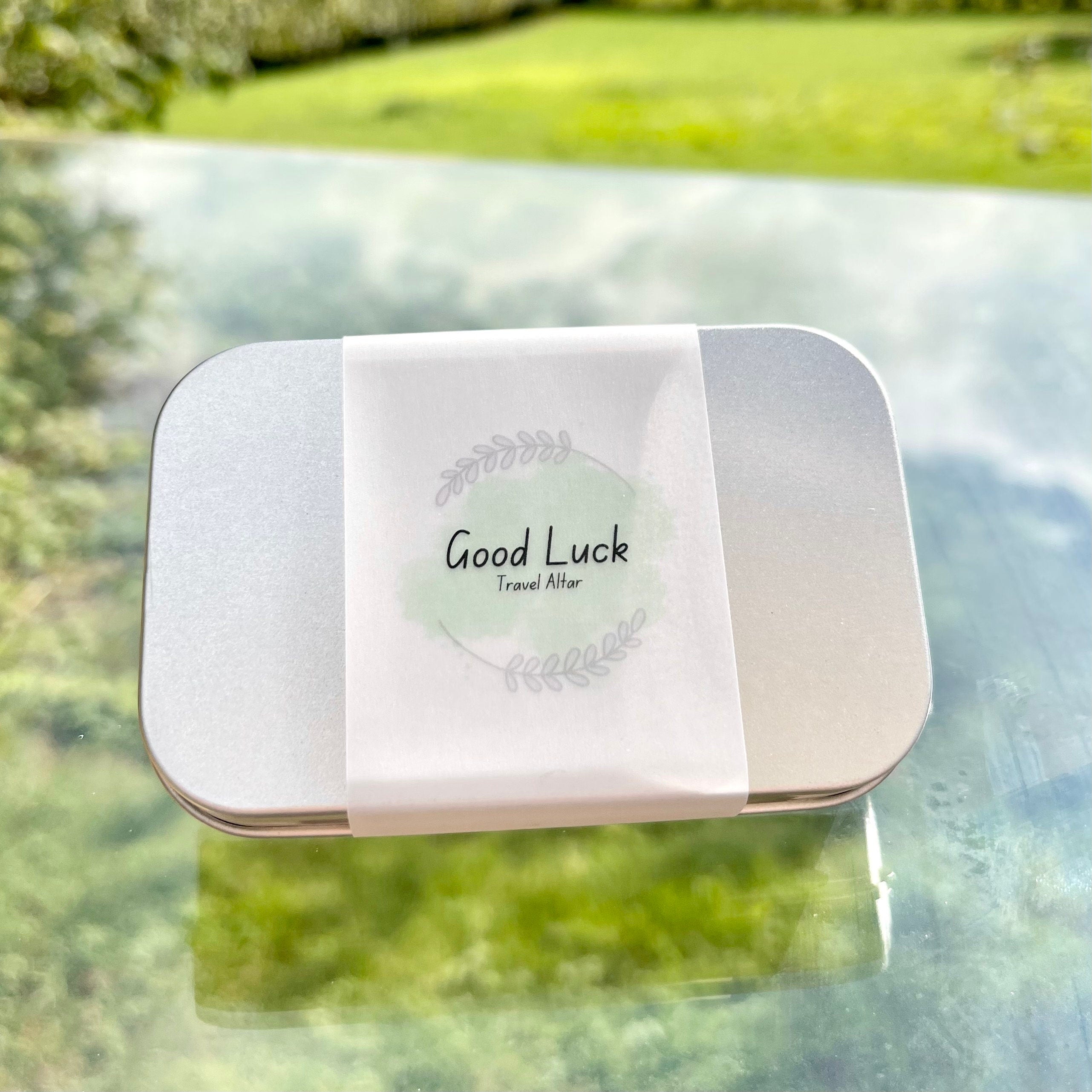 A metal tin with paper wrap with a green good luck travel altar logo on it