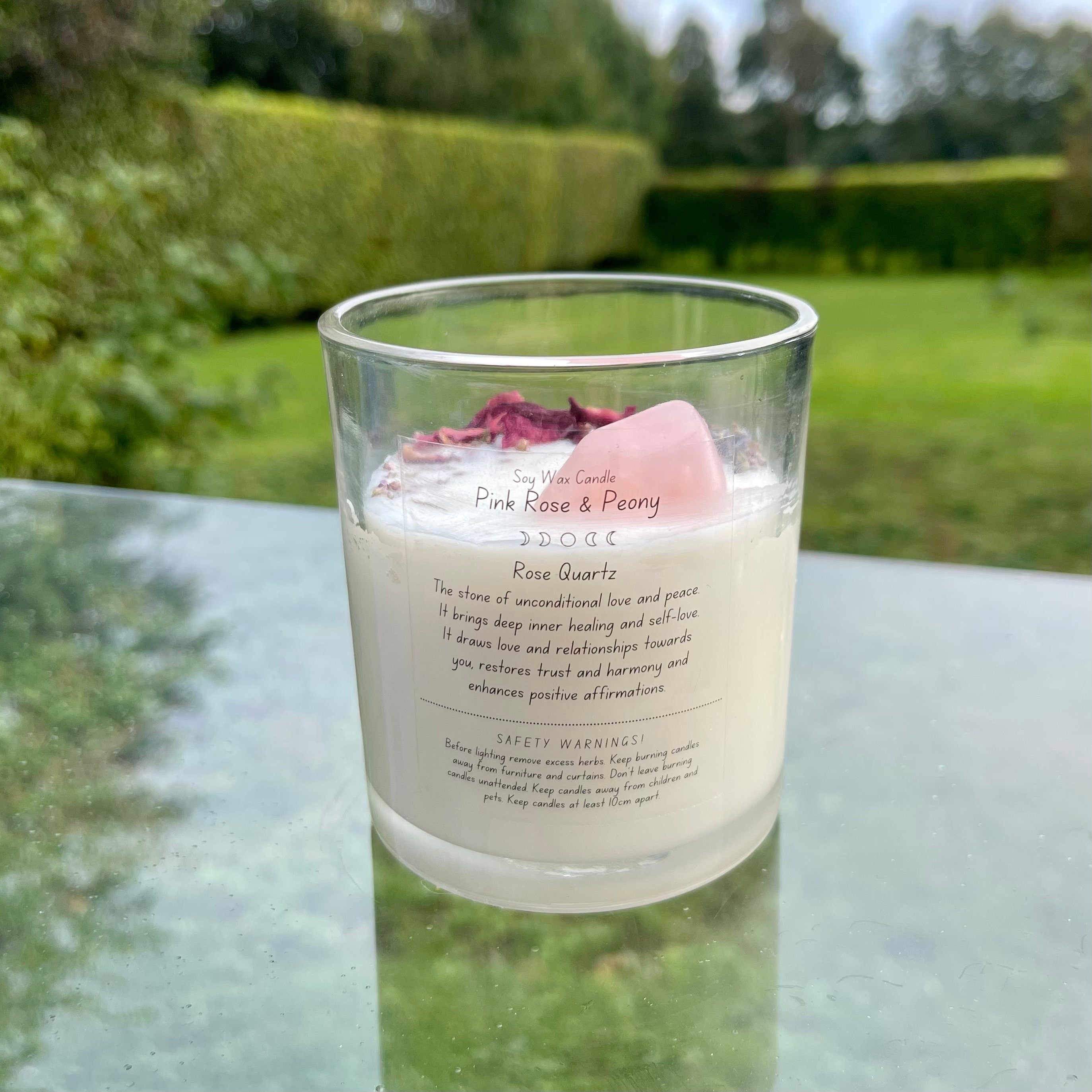A glass jar with white soy wax inside with a rose quartz tumblestone and rose petals set in the top. The back of the candle has a label explaining the crystal and herbs