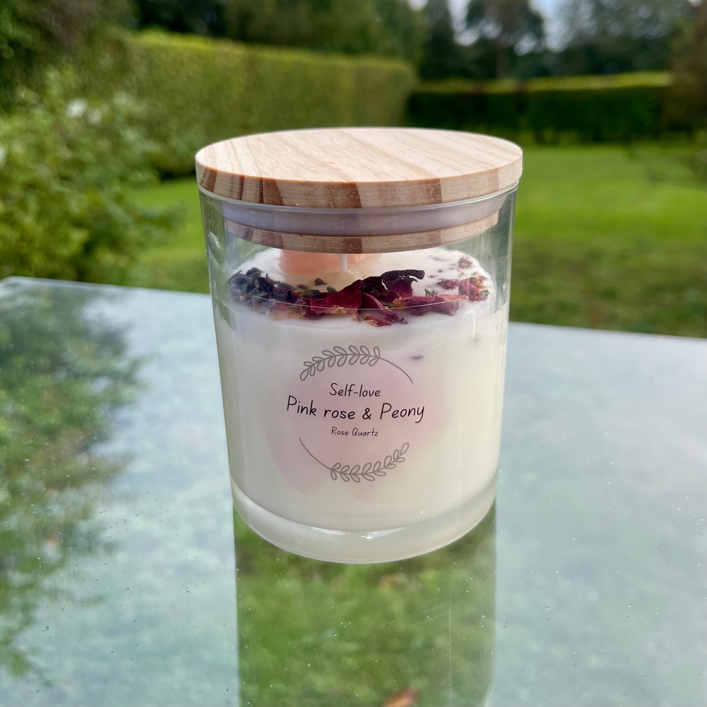 A glass jar with white soy wax inside with a rose quartz tumblestone and rose petals set in the top. the jar has a bamboo lid.