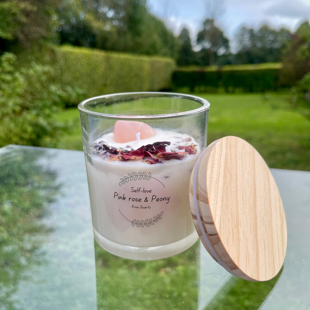 A clear glass jar with a wooden lid containing a white soy wax candle with rose petals and a pink rose quartz crystal on top. The candle is set in an outdoor environment with greenery in the background.