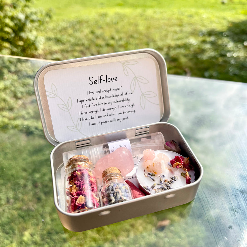 A travel altar kit for self-love, opened to display its contents including a spell candle, rose quartz stone, jars of dried rose petals and lavender, and a rose scented incense cone, all arranged neatly inside a small tin box with the words 'Self Love' written on the lid.