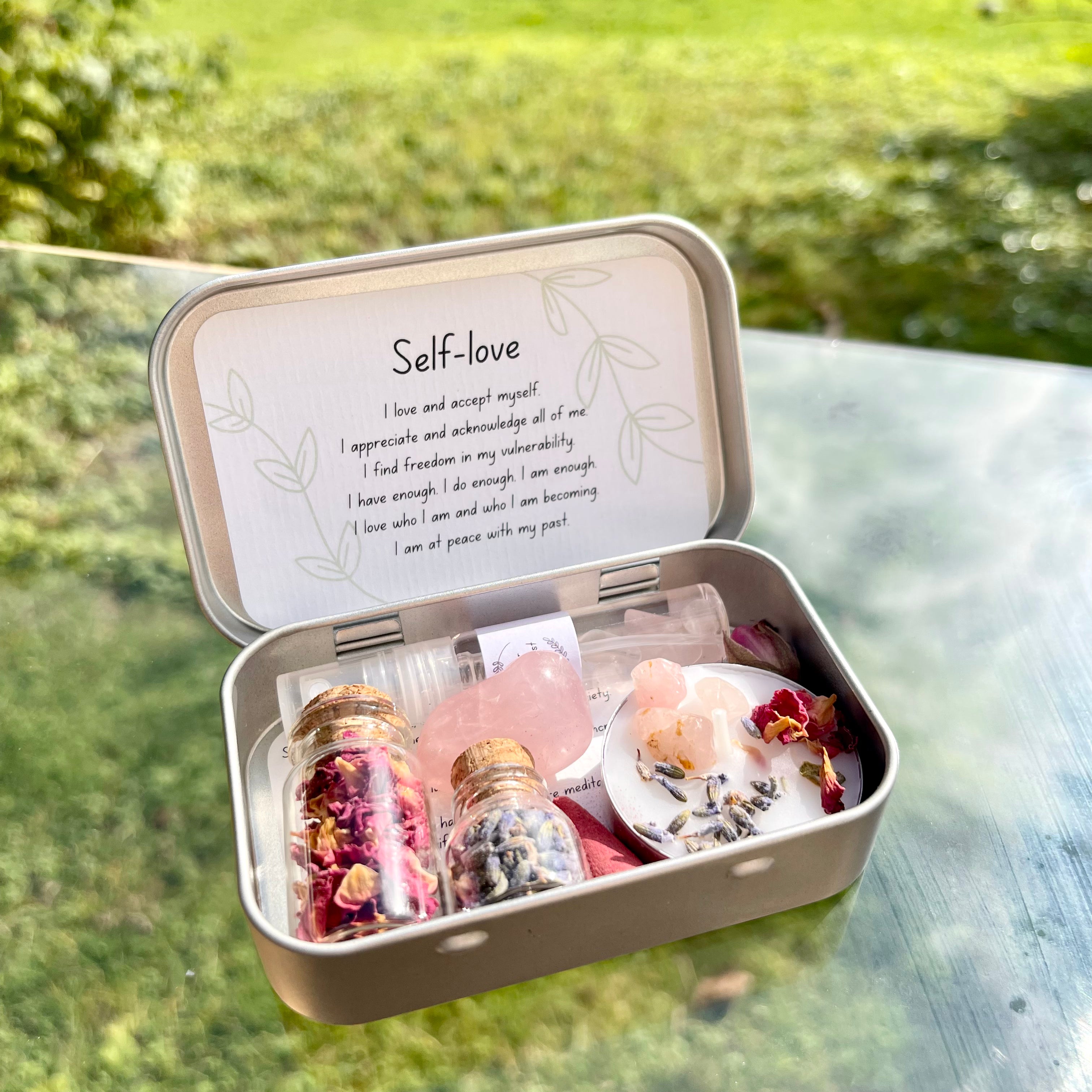 A travel altar kit for self-love, opened to display its contents including a spell candle, rose quartz stone, jars of dried rose petals and lavender, and a rose scented incense cone, all arranged neatly inside a small tin box with the words 'Self Love' written on the lid.