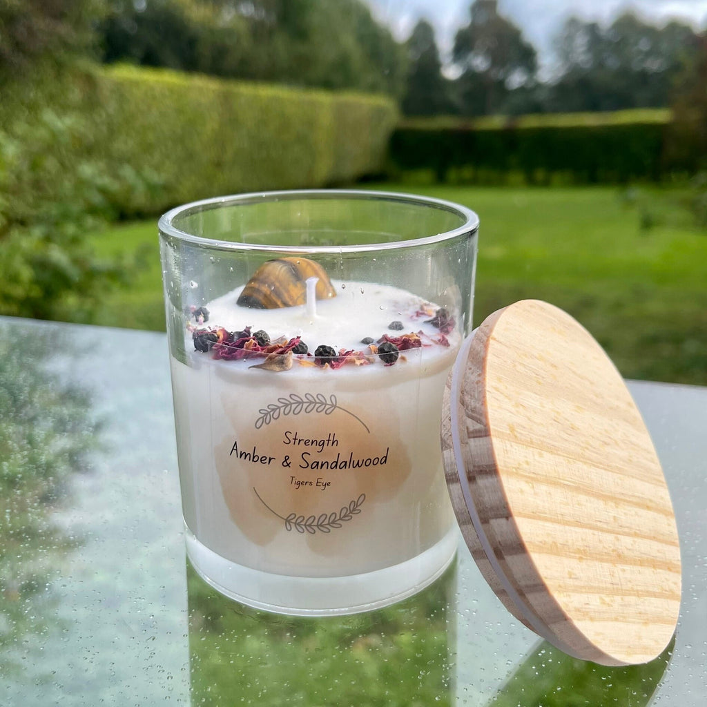 A soy wax candle with amber and sandalwood scent, topped with a tiger's eye crystal and herbs, in a clear glass jar with a wooden lid.