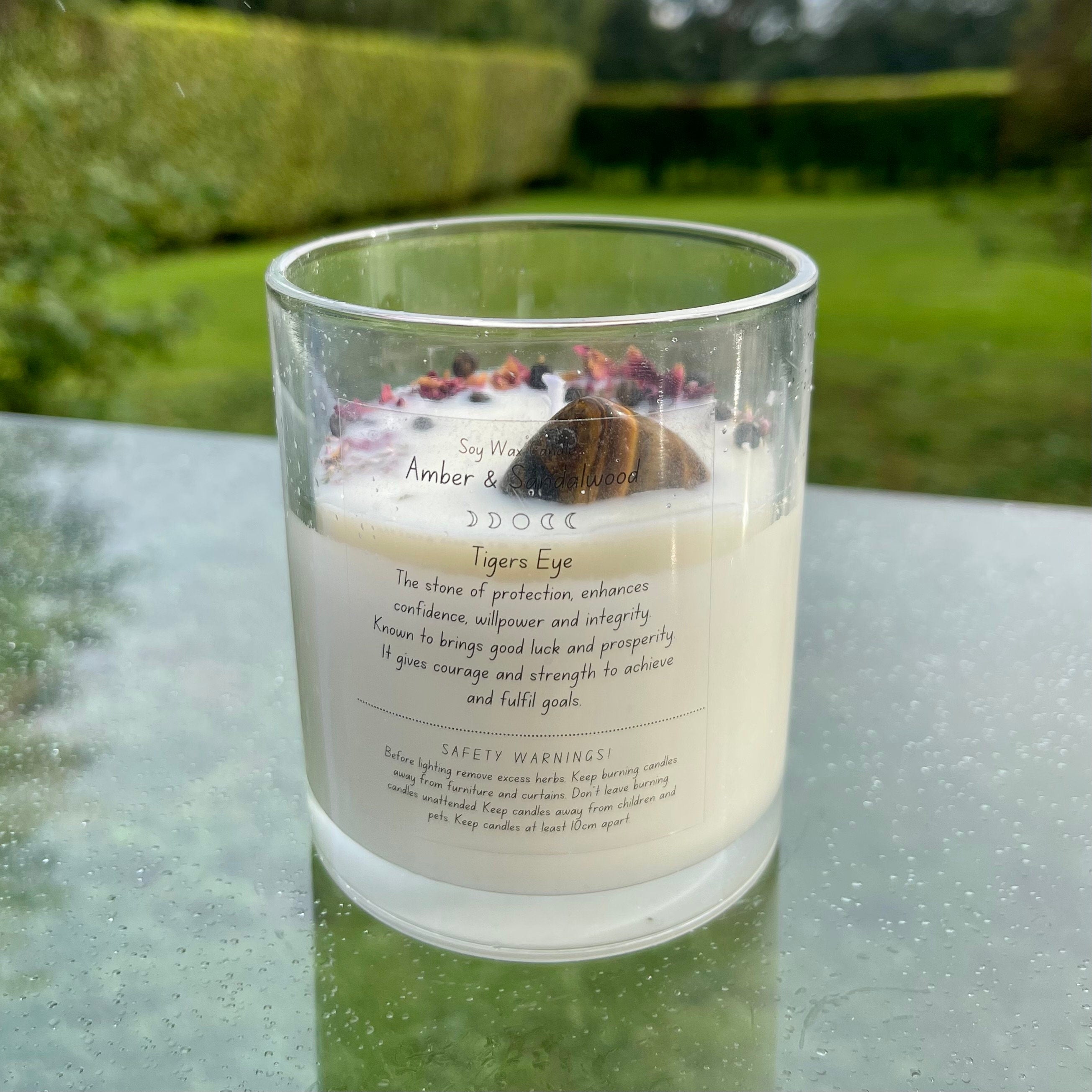 A glass jar with white soy wax inside with a tigers eye tumblestone and herbs set in the top. The back of the candle has a label explaining the crystal and herbs