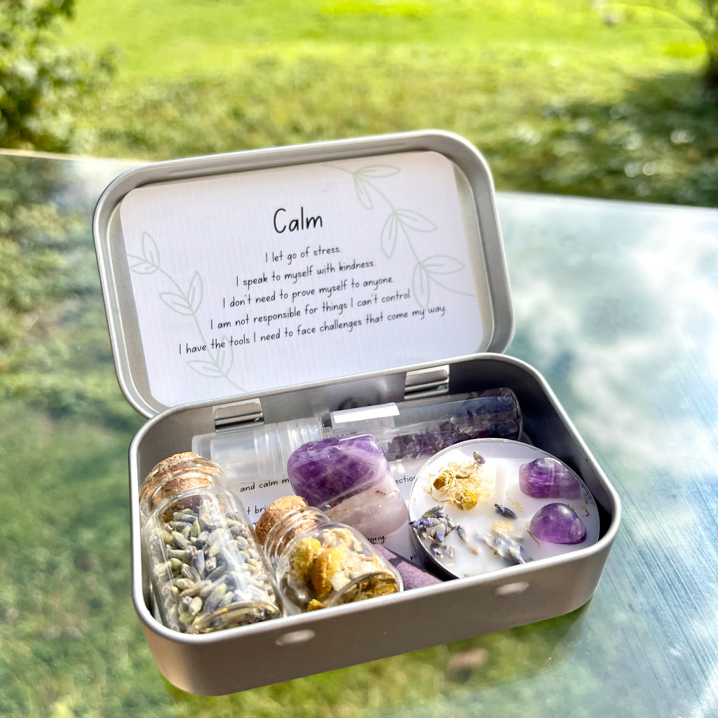 A portable travel altar tin opened to display various items for meditation and spell crafting, including a spell candle, jars of dried herbs, and an amethyst stone,and incense, set against an outdoor background.