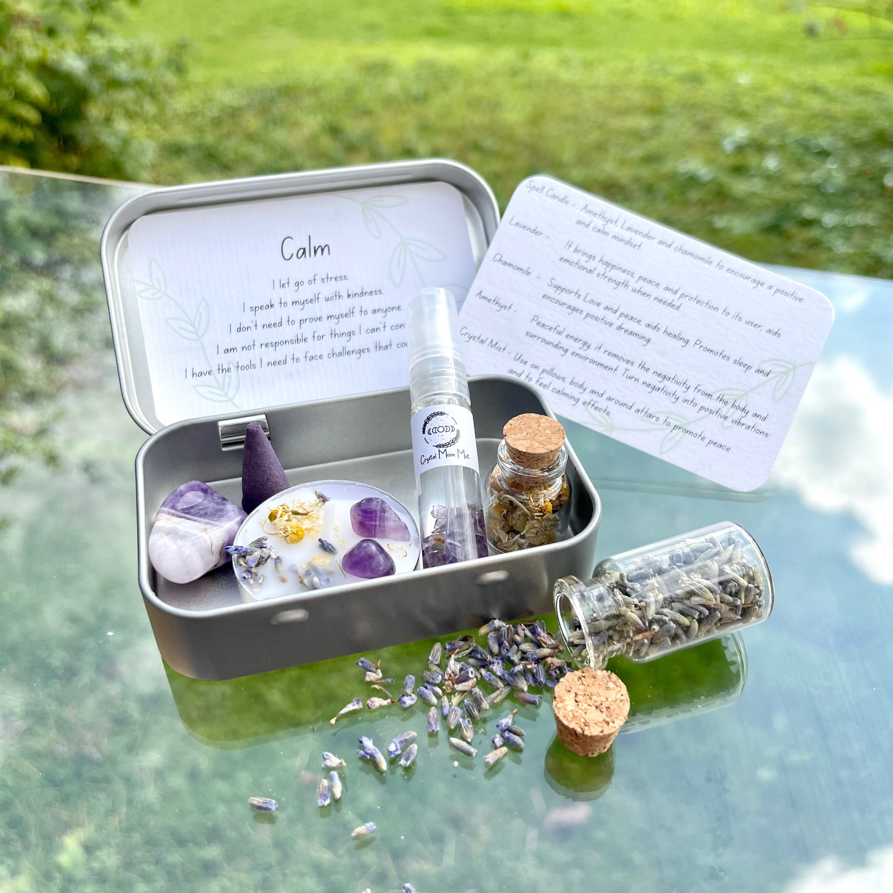 A portable calm themed travel altar tin opened to display its contents including a white spell candle with lavender petals, chamomile, and amethyst stone, alongside jars of chamomile and lavender, and a crystal infused lavender moon water bottle. The altar tin is placed on a surface with scattered lavender petals around it.
