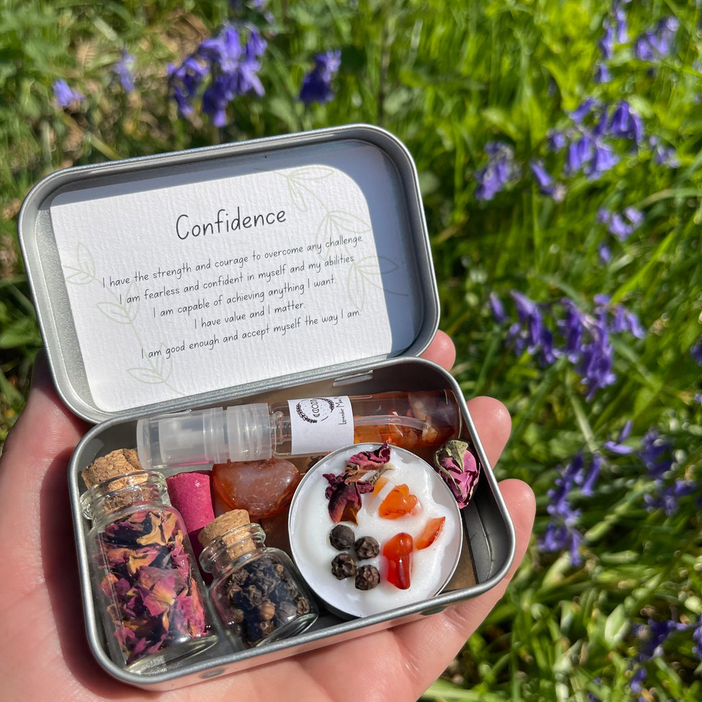 A confidence travel altar held in a hand, the tin is open and shows the contents neatly arranged. Inside is a moon mist, 2 jars of herb, a carnelian tumblestone, a spell candle and incense cone.