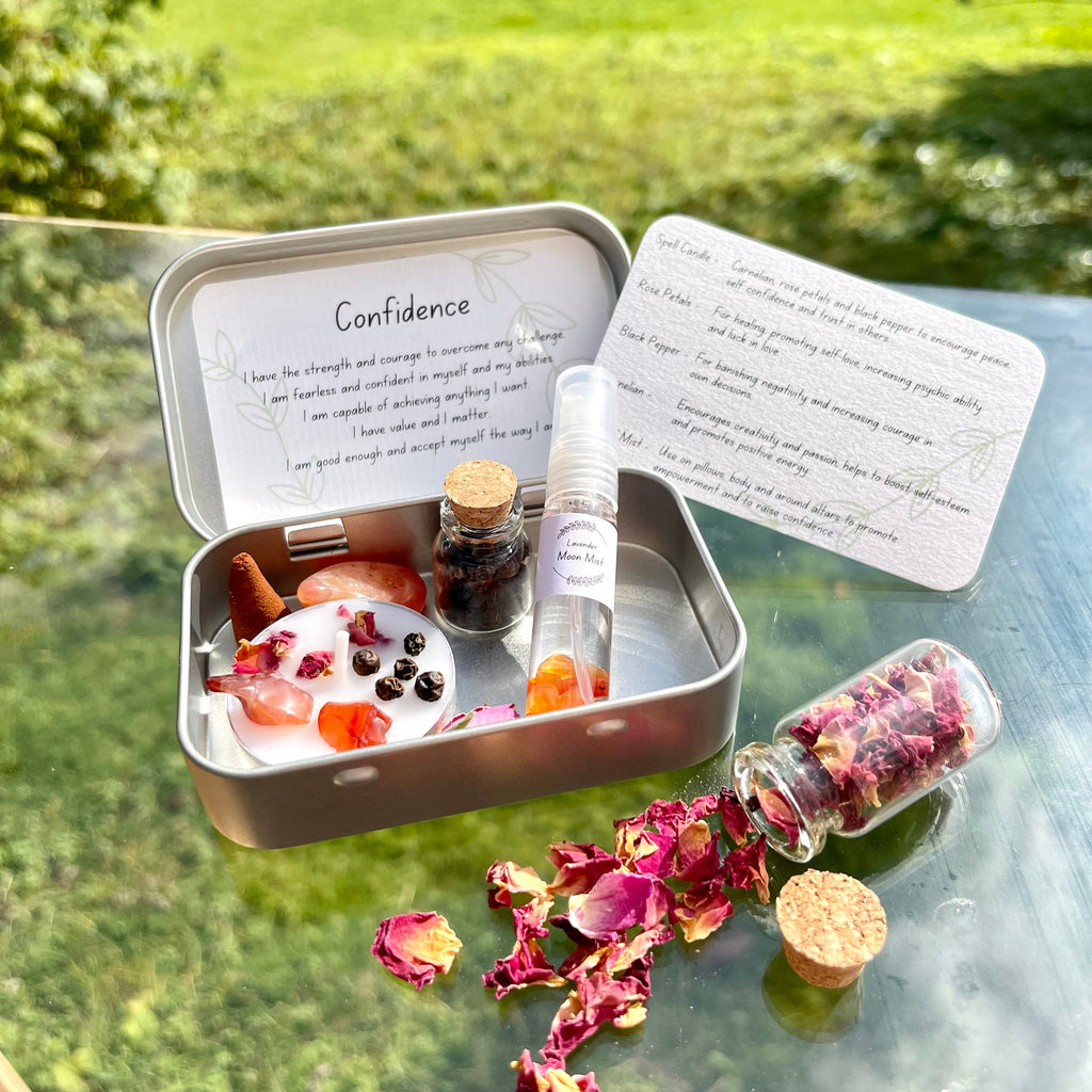 A portable Confidence themed travel altar tin opened to display its contents including a white spell candle with rose petals, black pepper, and carnelian stone, alongside jars of black pepper and rose petals, and a crystal infused lavender moon water bottle. The altar tin is placed on a surface with scattered rose petals around it.