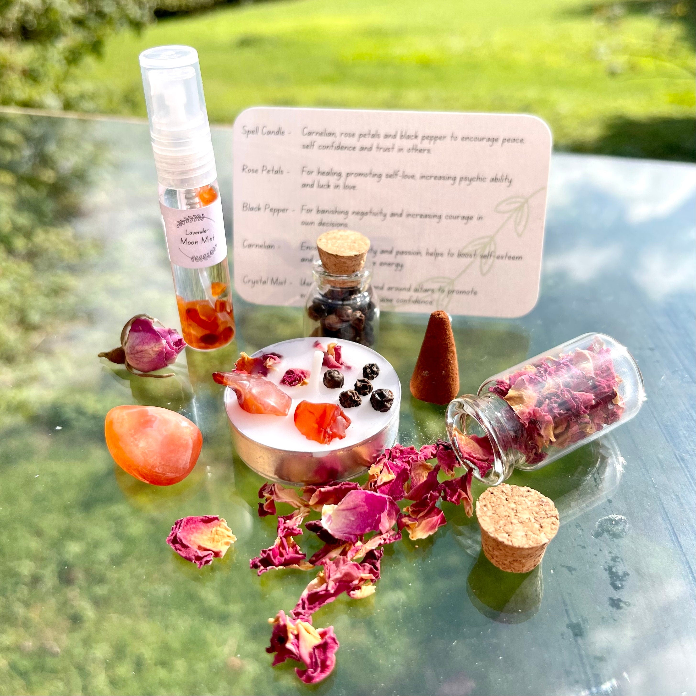 The contents of the confidence travel altar laid out. A moon mist with carnelian crystal chips, a jar of rose petals, jar of pepper, spell candle, incense cone and carnelian tumblestone.