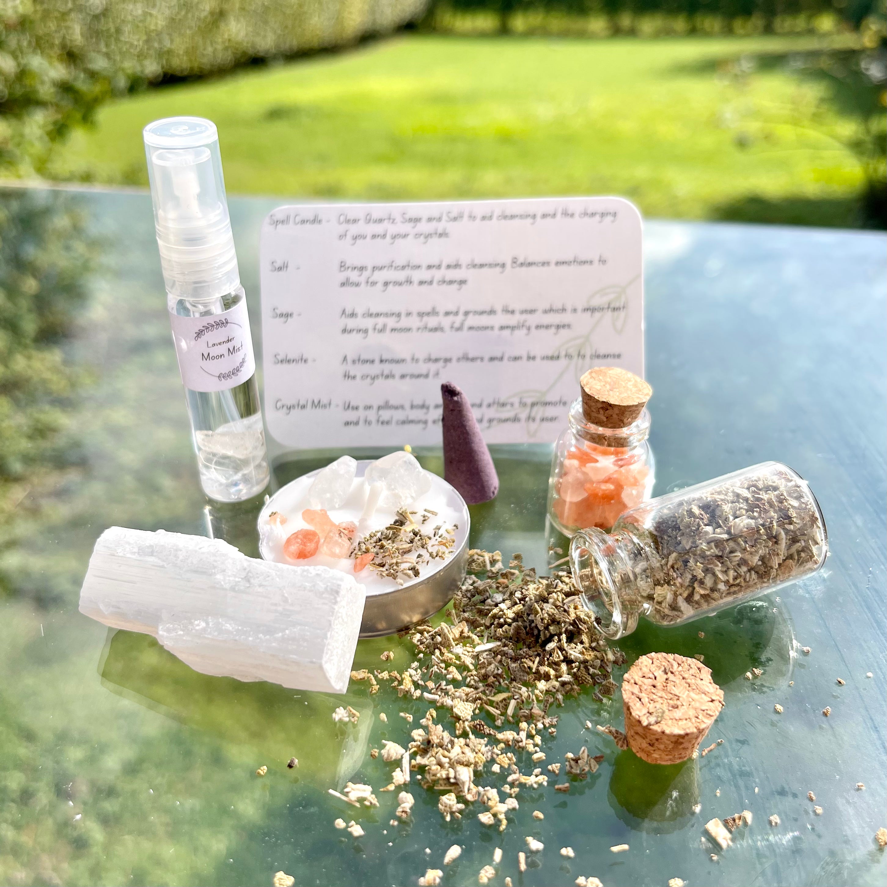 The contents of the full moon cleansing travel altar laid out. A moon mist with clear quartz crystal chips, a jar of sage, jar of salt, spell candle, incense cone and selenite stick.