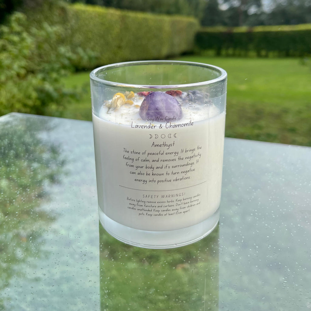 A glass jar with white soy wax inside with an amethyst tumblestone and herbs set in the top. The back of the candle has a label explaining the crystal and herbs