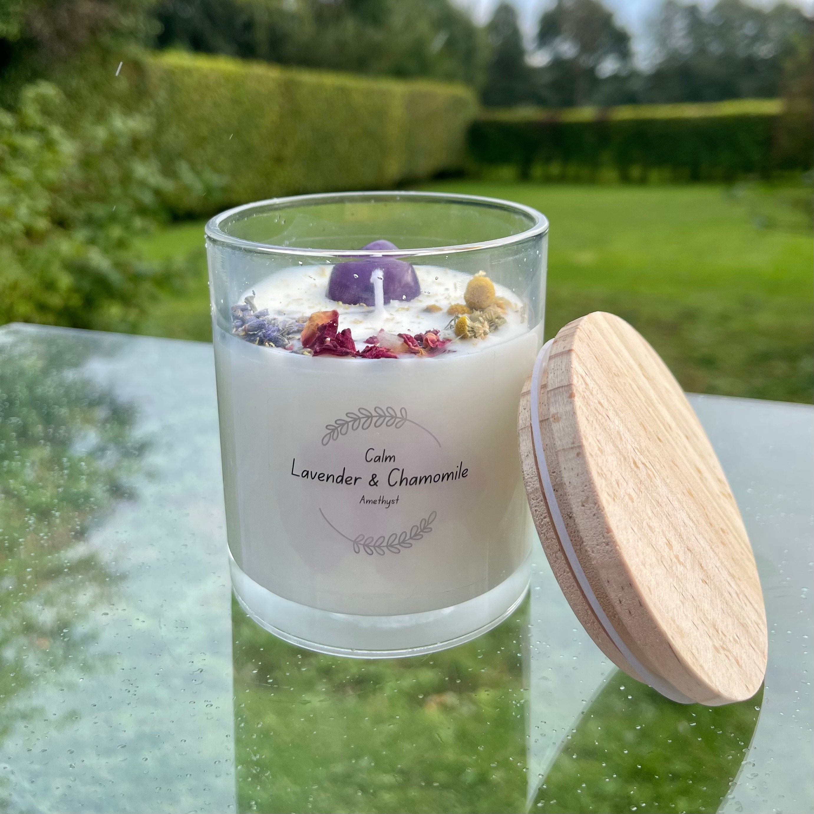 A clear glass Soy Wax candle with lavender and chamomile essential oils, adorned with a purple Amethyst Crystal and floral petals, with a wooden lid beside it.
