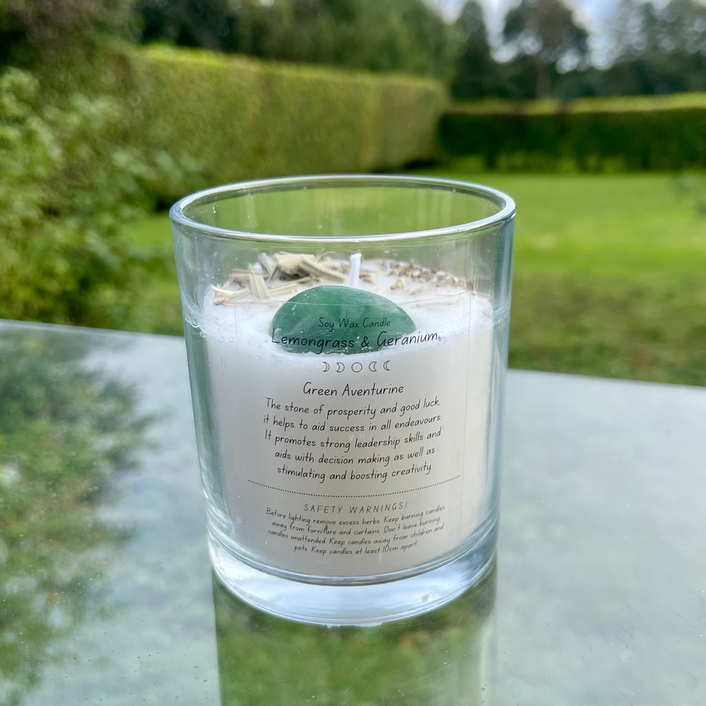 A glass jar with white soy wax inside with an aventurine tumblestone and herbs set in the top. The back of the candle has a label explaining the crystal and herbs