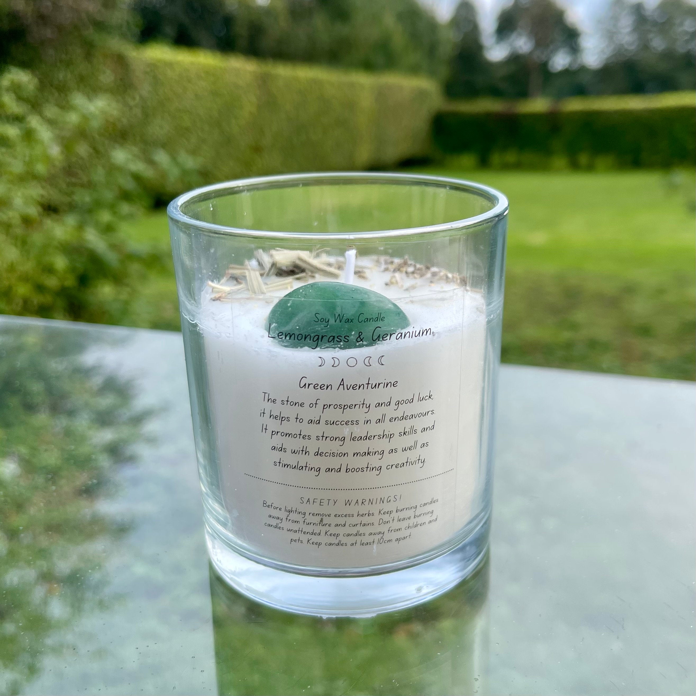 A glass jar with white soy wax inside with an aventurine tumblestone and herbs set in the top. The back of the candle has a label explaining the crystal and herbs