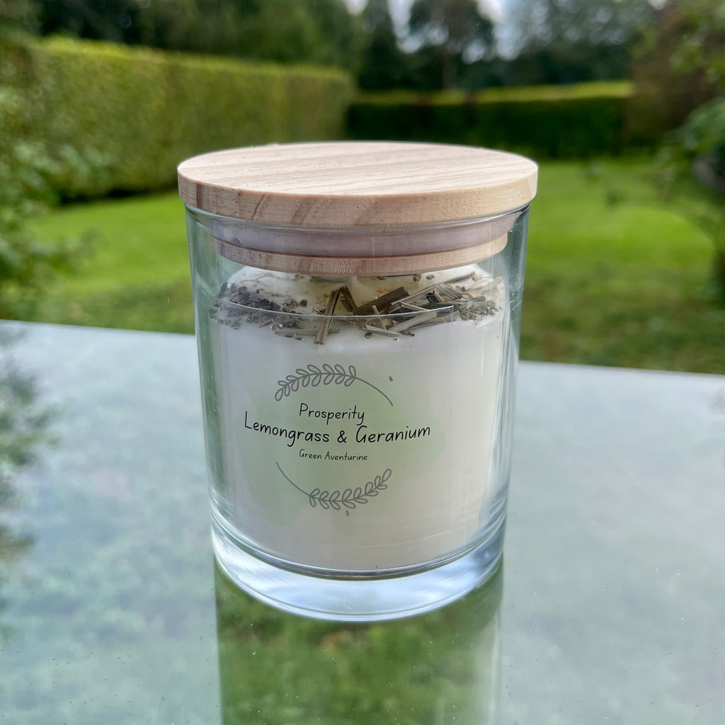 A glass jar with white soy wax inside with a green aventurine tumblestone and rose petals set in the top. the jar has a bamboo lid.