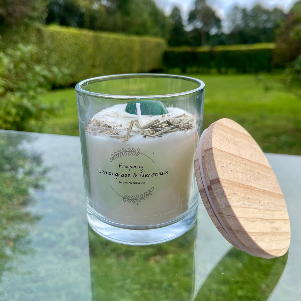 A clear glass soy wax candle with lemongrass and geranium essential oils, featuring a wooden lid and a green aventurine crystal on top with herbs, set on a glass surface with a natural background.