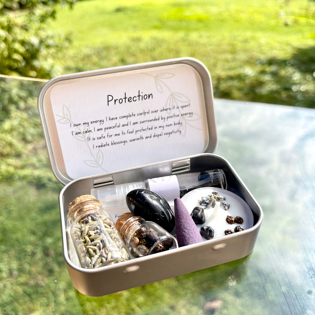 A travel altar kit for protection, opened to display its contents including a spell candle, black tourmaline, jars of dried lavender petals and pepper, and a lavender scented incense cone, all arranged neatly inside a small tin box with the words 'protection' written on the lid.