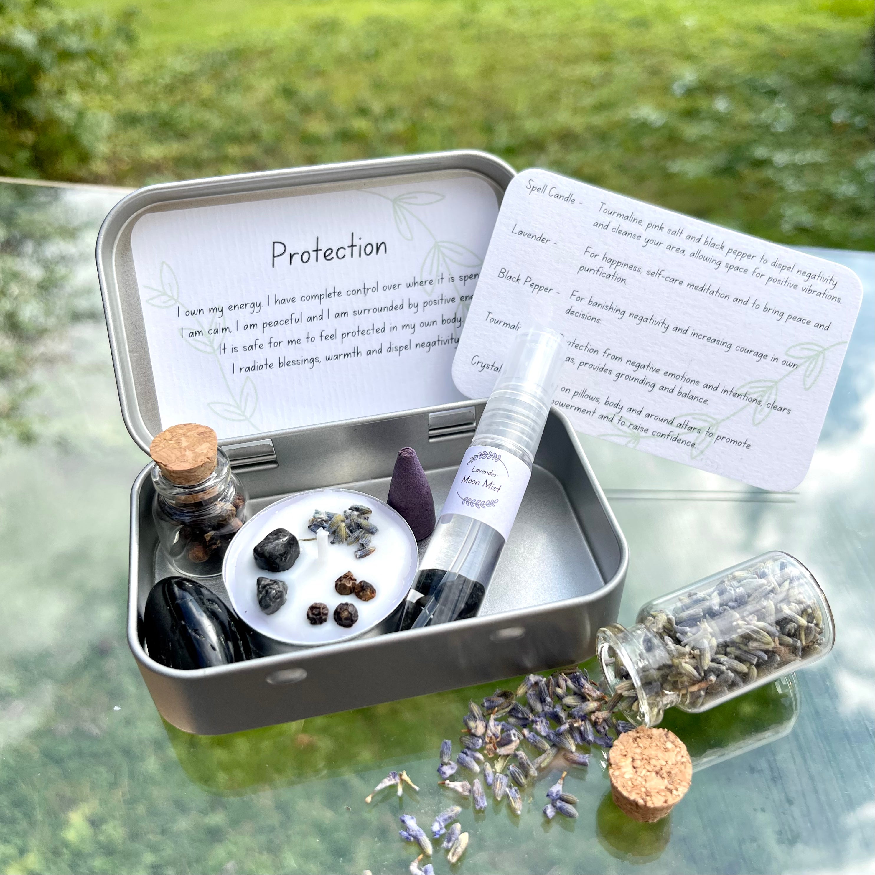 A portable protection themed travel altar tin opened to display its contents including a white spell candle with lavender petals,pepper, and tourmaline stone, alongside jars of pepper and lavender, and a crystal infused lavender moon water bottle. The altar tin is placed on a surface with scattered lavender petals around it.