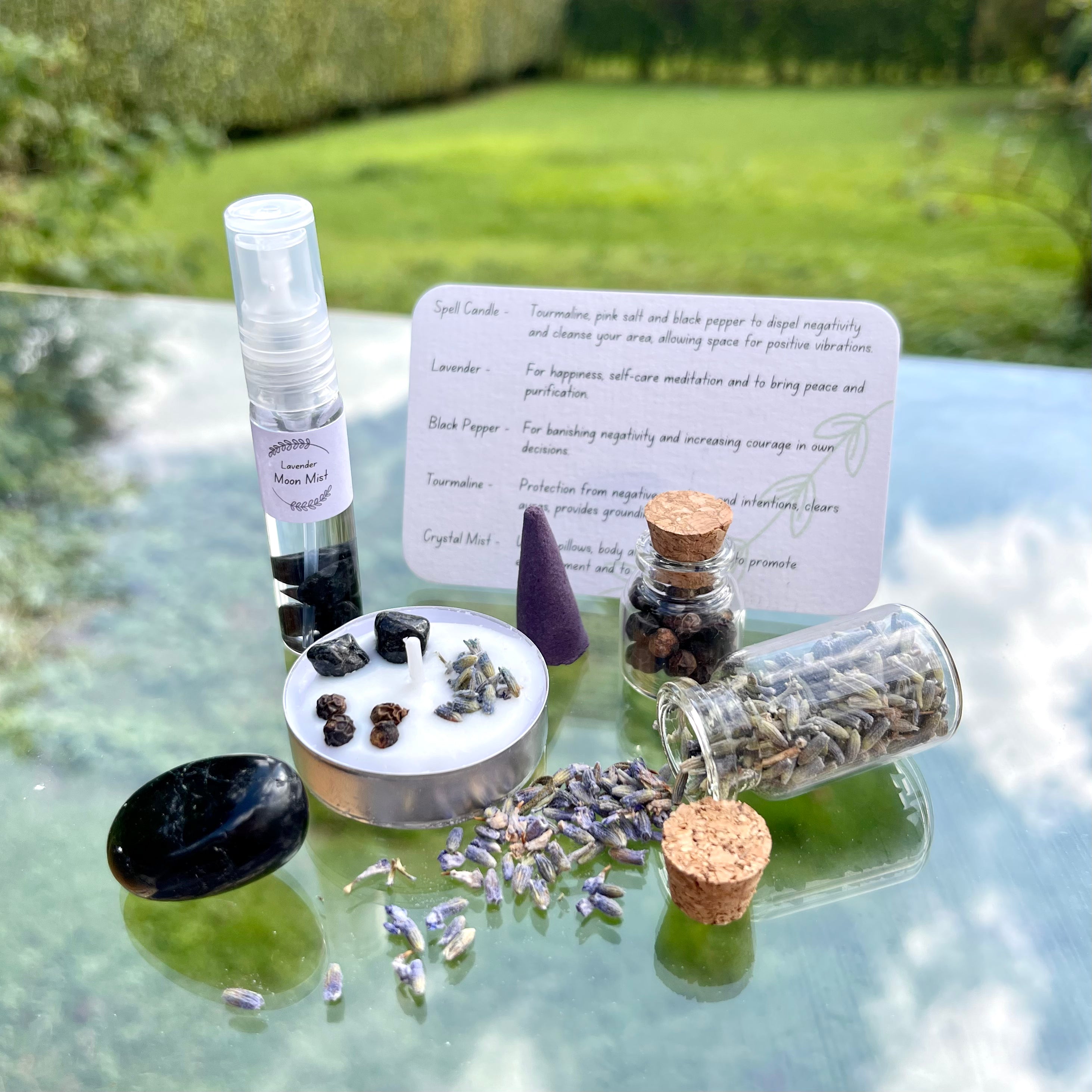 The contents of the protection travel altar laid out. A moon mist with tourmaline crystal chips, a jar of pepper, jar of lavender, spell candle, incense cone and tourmaline tumblestone.