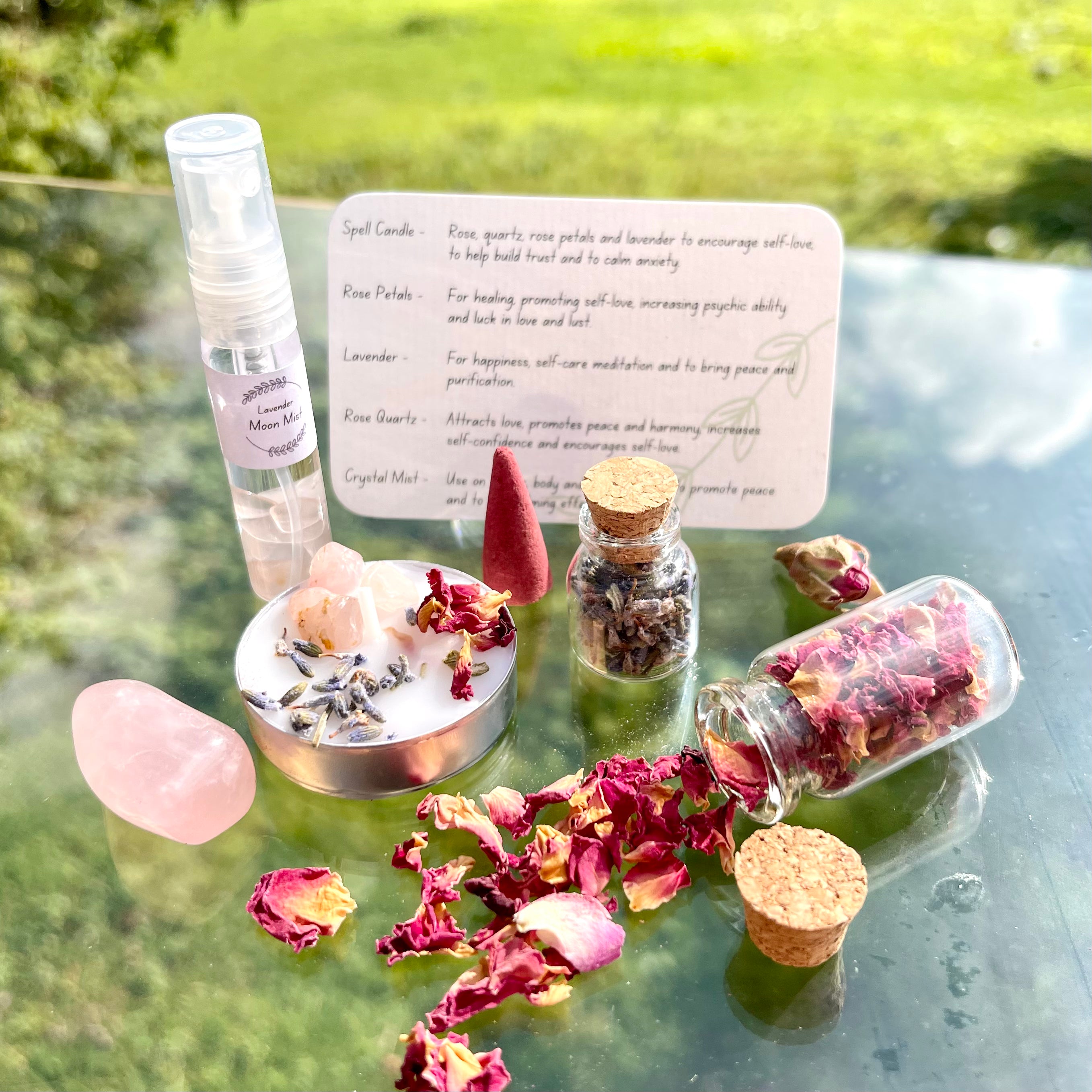 The contents of the self love travel altar laid out. A moon mist with rose quartz crystal chips, a jar of rose petals, jar of lavender, spell candle, incense cone and rose quartz tumblestone.
